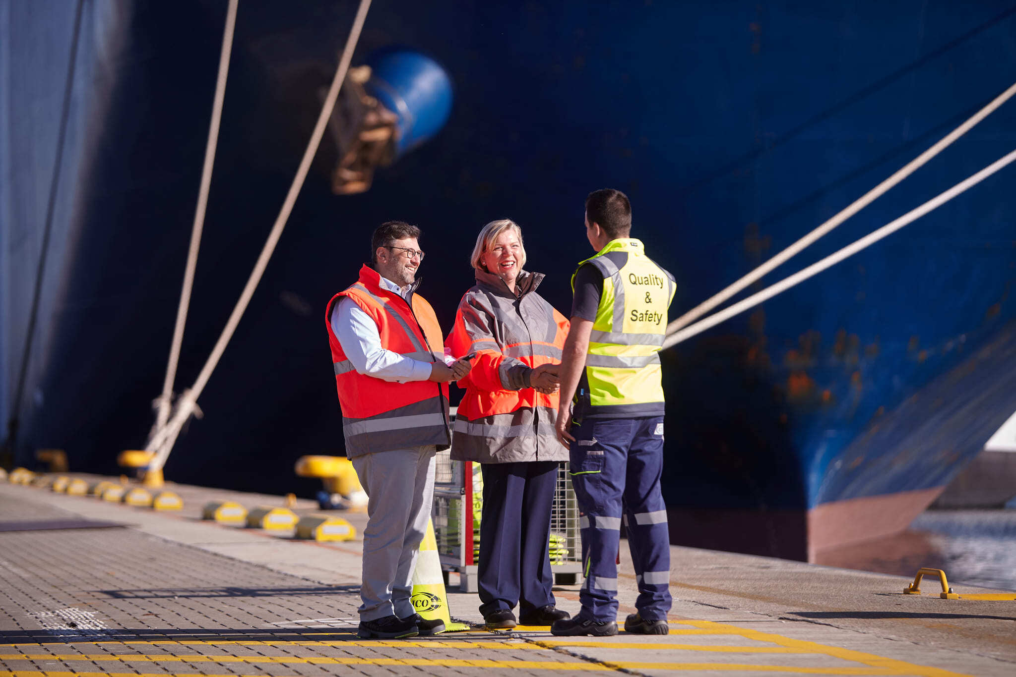 Drie havenmedewerkers in fluovesten geven elkaar de hand voor een groot schip met opschrift Quality & Safety